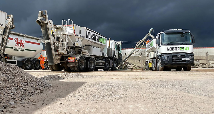 construction site featuring several concrete mixer trucks under a dark, cloudy sky. In the foreground, there's a pile of aggregate material.