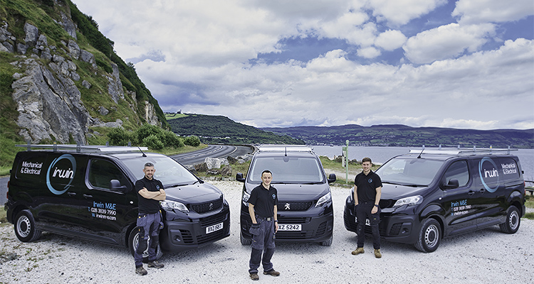 three individuals standing in front of three black vans with the "Irwin M&E" logo, set against a scenic backdrop of mountains and a body of water