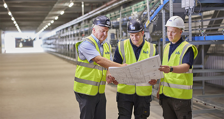 three men wearing safety vests and hard hats, gathered around a large blueprint or set of plans within a warehouse
