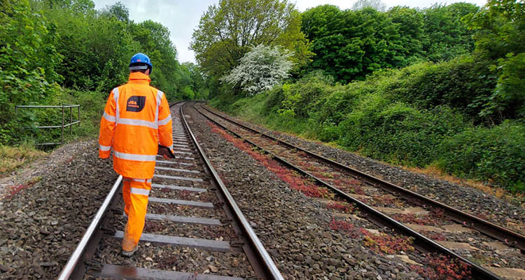 a railway worker in high-visibility orange personal protective equipment walking along a set of train tracks