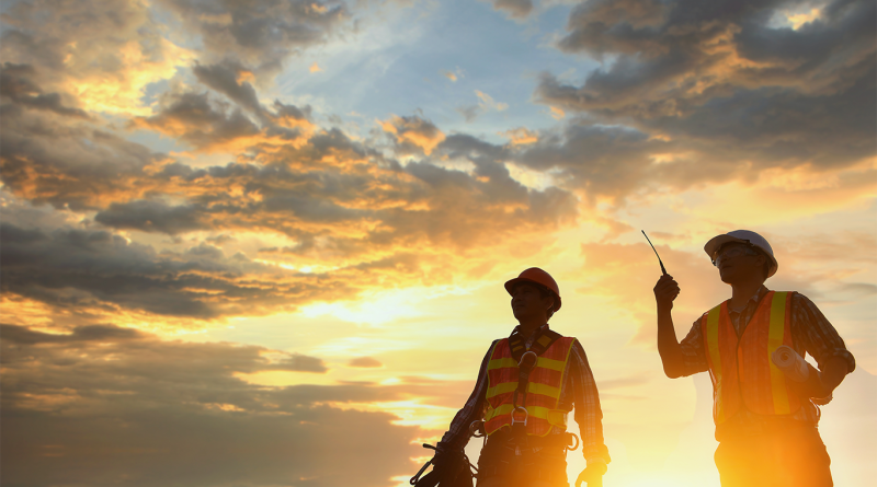 Silhouetted construction workers against a sunset sky. One worker carries a rope while the other holds a walkie-talkie and building plans.